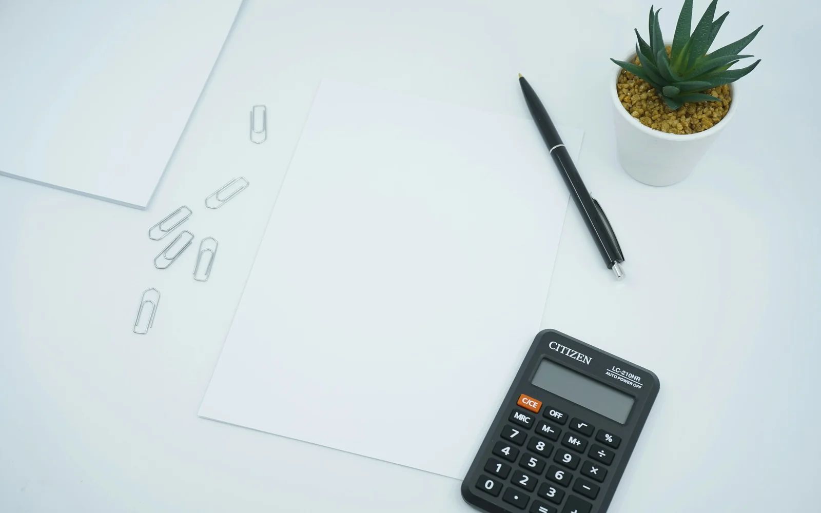 A calculator, pen and blank sheets of paper on a pale desk.