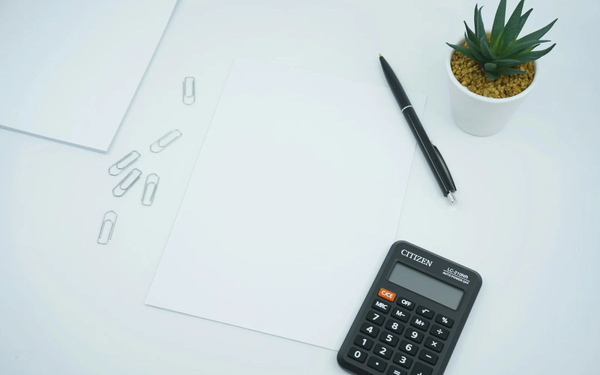 A calculator, pen and blank sheets of paper on a pale desk.