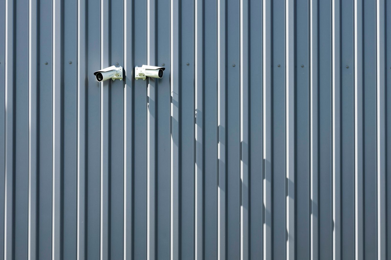Two white surveillance cameras casting long shadows on a blue-gray corrugated wall.