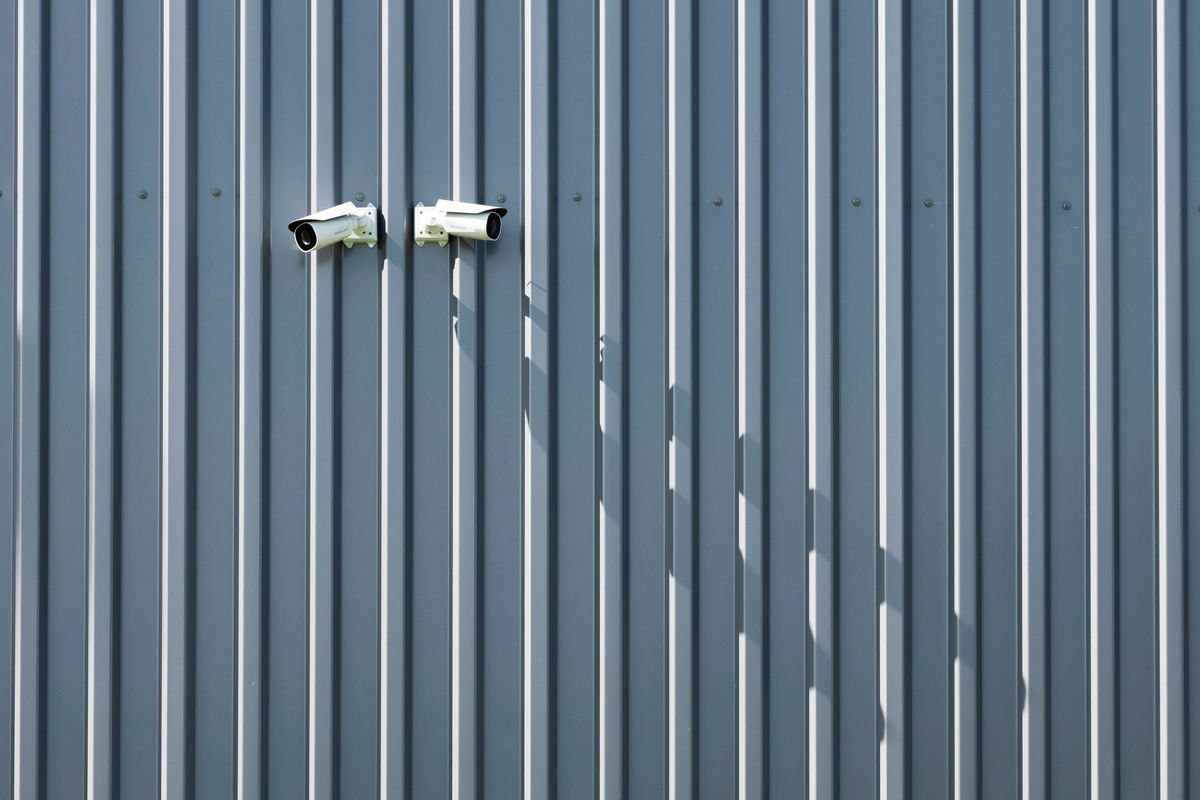 Two white surveillance cameras casting long shadows on a blue-gray corrugated wall.